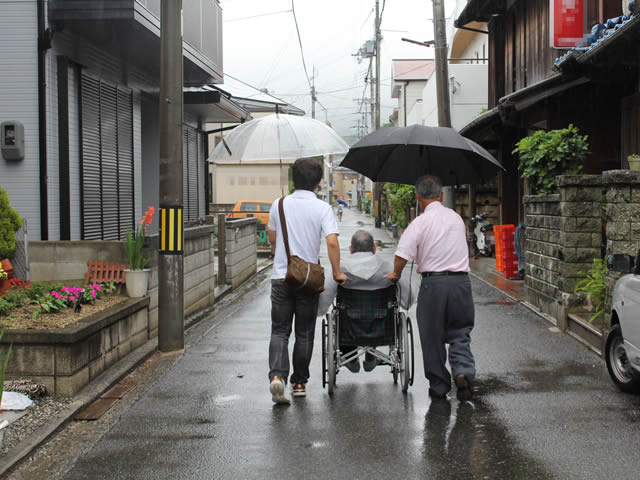 写真5：車椅子を押され、傘をさして、道幅３～４メートルほどの地区のメインストリートを歩く写真です。