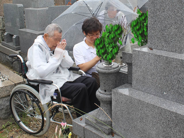 写真6：車椅子に乗り、お墓の前で手を合わせるお客様とエスコートヘルパーの写真です。